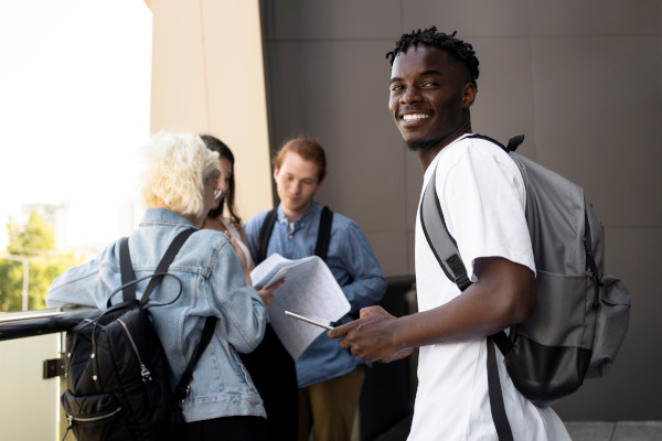 Foto: grupo de estudantes reunidos no corredor de um bloco da universidade; em primeiro plano, um estudante negro de cabelos curtos e pretos, com camiseta branca e mochila cinza.