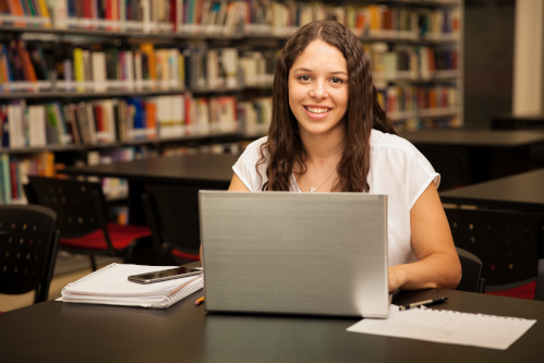 Foto: Estudante, mulher, jovem, de cabelos longos, ondulados, castanhos, vestindo camiseta branca, usa notebook em biblioteca 