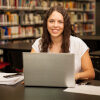 Foto: Estudante, mulher, jovem, de cabelos longos, ondulados, castanhos, vestindo camiseta branca, usa notebook em biblioteca