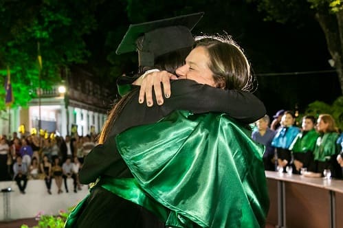 Foto de professora com veste talar e adereços na cor verde abraçando, durante cerimônia de colação de grau, uma formanda vestindo beca e faixa verde.