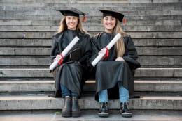 Fotografia quadrada: duas jovens concludentes sentadas vestem beca e seguram seus diplomas físicos