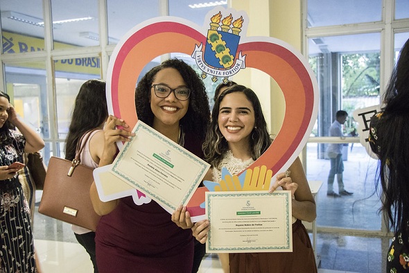 Fotografia: duas estudantes seguram seus certificados de distinção acadêmica e uma moldura de coração com o brasão da UFC