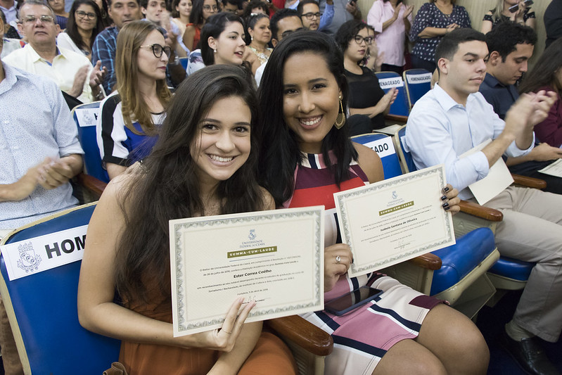 Duas egressas sorriem ao exibir seus diplomas com distição acadêmica summa cum laude