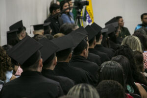 #PraCegoVerFoto de uma fileira de estudantes de costas e com capelo na cabeça 